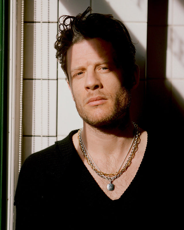 James Norton wearing a black top and Stephen Einhorn gold and silver chain necklace against a tiled wall.